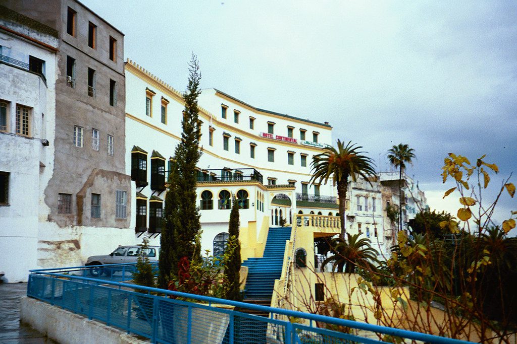 View of the historic old city of Tangier, showcasing narrow winding streets lined with whitewashed buildings, vibrant blue doors, and colorful flower pots. The image captures the charming blend of Moroccan architecture and coastal ambiance, with glimpses of the nearby Mediterranean Sea in the background