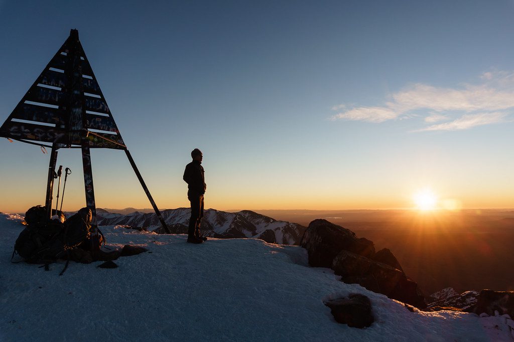 View from the summit of Toubkal mountain, showing rocky peaks and snow patches with clear skies above. The vast Moroccan landscape stretches out below, revealing valleys and distant ridgelines