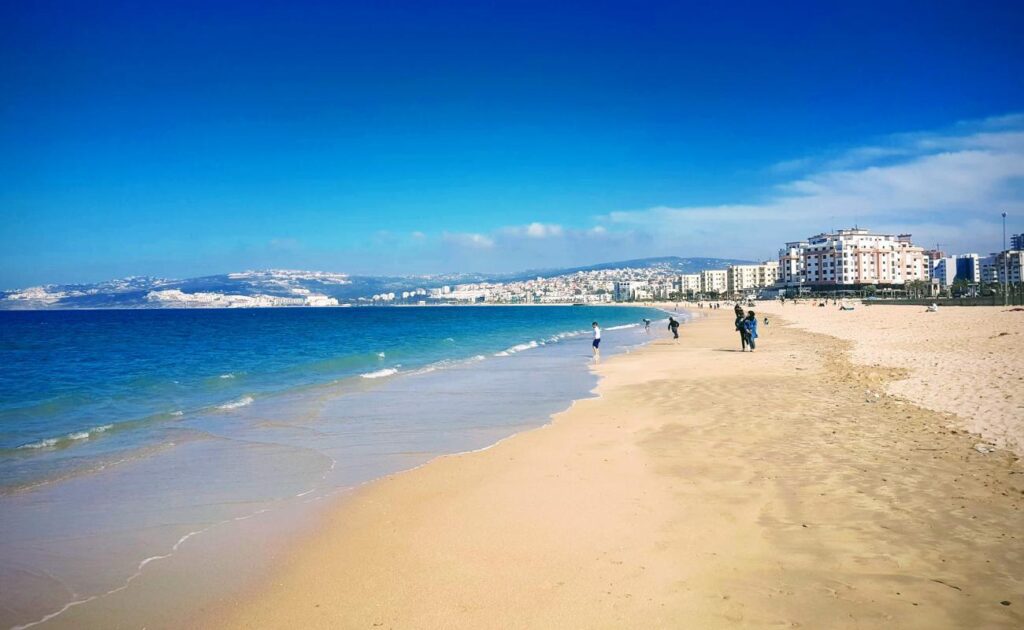Panoramic view of Plage de Tanger, featuring golden sandy shores lined with beach umbrellas and sunbathers. The turquoise waters of the Mediterranean Sea gently lap at the beach, with the picturesque Tangier skyline and coastal hills visible in the background under a bright blue sky.