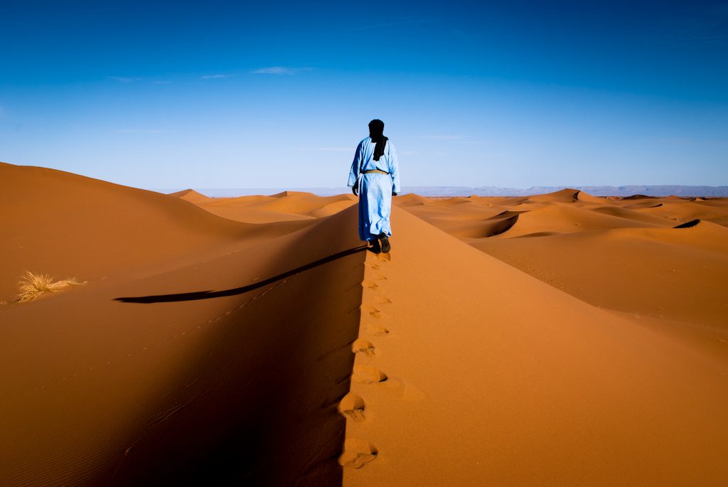 Golden sand dunes of the Moroccan Sahara under a clear blue sky with footprints leading into the distance.