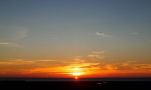 Stunning sunset over Achakar Beach in Tangier, with vibrant hues of orange, pink, and purple reflecting off the tranquil sea. Silhouetted palm trees line the sandy shore, creating a picturesque scene as waves gently lap against the beach