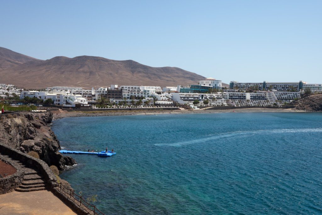 Stunning view of Playa Blanca beach in Tangier, featuring soft white sands and crystal-clear turquoise waters. The beach is lined with sun loungers and umbrellas, with palm trees swaying in the gentle breeze. In the background, the rugged cliffs and the vibrant blue sky create a picturesque coastal scene.