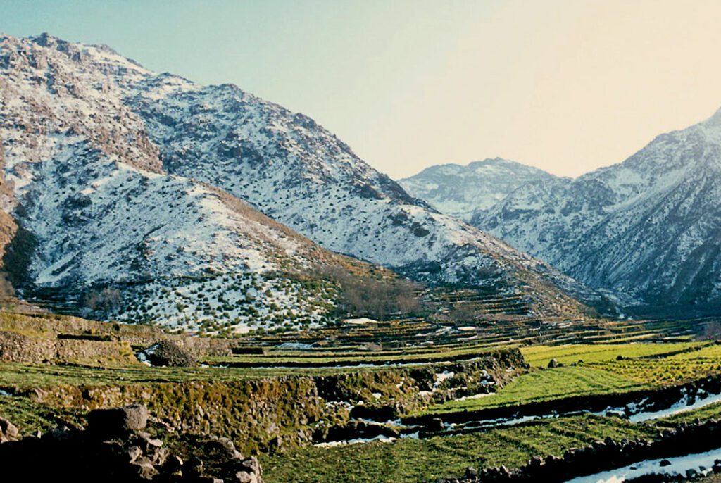 Scenic view of Imlil Valley, featuring lush green terraced fields, traditional Berber houses, and the surrounding Atlas Mountains. The landscape is bathed in sunlight, showcasing the vibrant colors of nature against the majestic mountain backdrop.