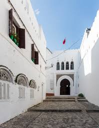 View of the historic old city of Tangier, showcasing narrow winding streets lined with whitewashed buildings, vibrant blue doors, and colorful flower pots. The image captures the charming blend of Moroccan architecture and coastal ambiance, with glimpses of the nearby Mediterranean Sea in the background