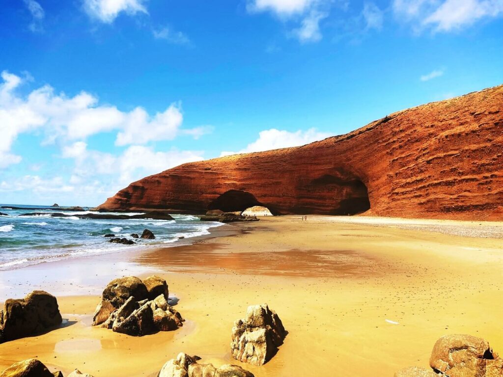 Stunning view of Legzira Beach, showcasing its iconic red cliffs, golden sand, and vibrant blue ocean under a clear sky.