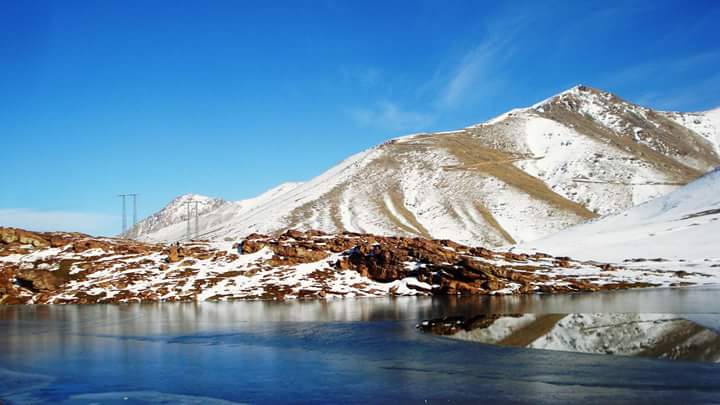 A panoramic view of Oukaimeden, a picturesque ski resort in the Atlas Mountains near Marrakech. The image features snow-covered slopes and pine trees under a bright blue sky, with the rugged peaks of the mountains in the background, showcasing a blend of winter sports and natural beauty.