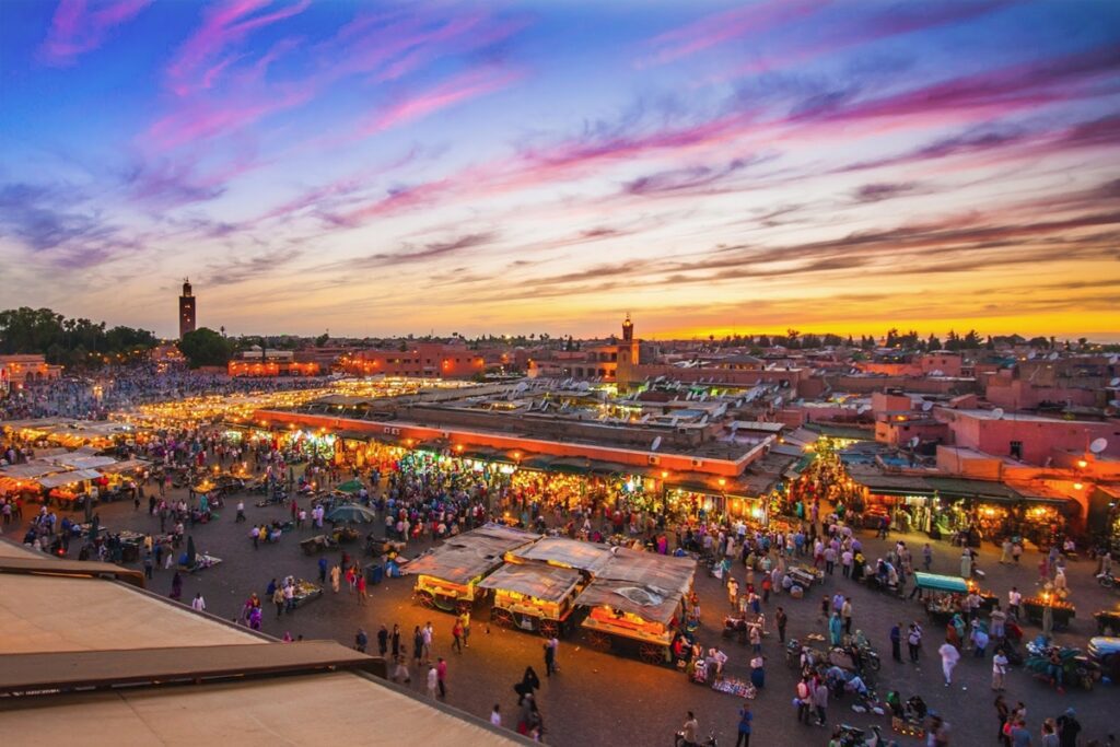 Vibrant scene at Jemaa el-Fnaa square in Marrakech, featuring bustling crowds, street performers, food stalls, and traditional Moroccan architecture. The sun sets behind the iconic Koutoubia Mosque, casting a warm glow over the lively atmosphere.