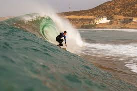 Surfers riding the waves at Taghazout beach, with golden sands and rocky cliffs in the background.