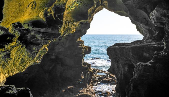 Panoramic view of the Caves of Hercules in Tangier, featuring dramatic limestone formations and the entrance shaped like a map of Africa. The azure waters of the Atlantic Ocean are visible in the background, with sunlight illuminating the cave's interior, showcasing its natural beauty and historical significance.