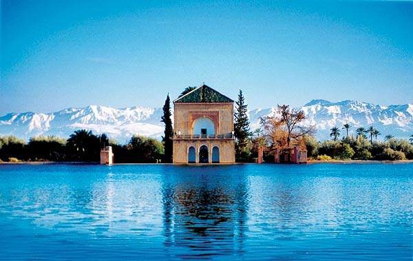 Scenic view of El-Menara, featuring the iconic pavilion surrounded by lush gardens and a large reflecting pool, with the majestic Atlas Mountains in the background under a clear blue sky.