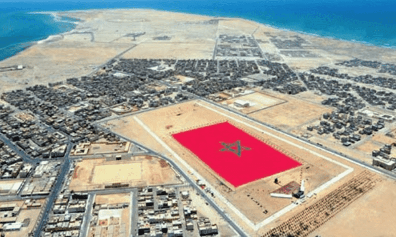 A panoramic view of Dakhla City, showcasing its vibrant coastal landscape, sandy beaches, and crystal-clear waters. The image captures colorful buildings lining the waterfront, with the backdrop of the desert and distant mountains, highlighting the city’s unique blend of natural beauty and urban charm.
